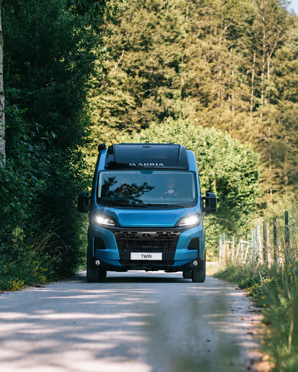 Der neue sturmblaue Fiat ADRIA Campervan fährt durch eine Waldstraße